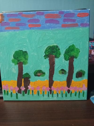 A flat canvas painting depicts a stylized landscape. The sky is a light blue with horizontal strokes of purple and coral, resembling abstract clouds. Below the sky is a solid band of turquoise, representing a field. In this field, three tall, brown-trunked trees with dense green foliage are spaced unevenly. Two smaller, green clumps of vegetation are also present in the field. A band of yellow runs along the bottom of the turquoise field, and below that, a line of small pink flowers with green stems is painted. The painting is placed on a dark wooden surface, possibly a desk or table. A keyboard is partially visible on the left side of the frame, and a cylindrical object, possibly a bottle, with a label reading "ROSE" is visible on the right. The background is a plain blue wall. The image is a photograph taken from a slightly elevated angle, looking down at the painting.