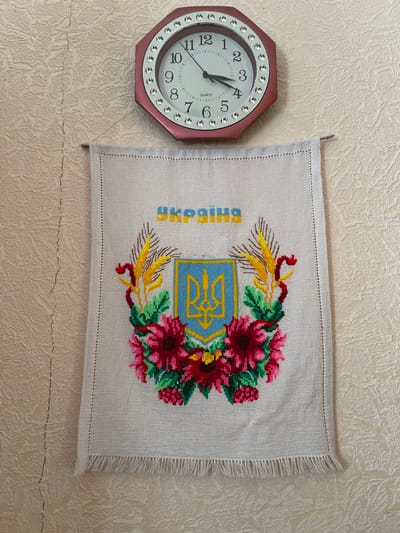 The scene depicts an interior wall in Pyriatyn, Ukraine. The wall is covered in light beige or off-white textured wallpaper, exhibiting a vertical crack on the left side. Two items are mounted on the wall. In the upper center, an octagonal wall clock with a pinkish-red frame is present, featuring clear, round decorative jewels around its perimeter. The clock has a white face with black Arabic numerals from 1 to 12. The hour hand points approximately halfway between 3 and 4, and the minute hand points directly at 4, indicating a time of 3:20. The word "QUARTZ" is printed below the center of the clock face. Directly beneath the clock, an embroidered textile hangs from a thin metal rod. The fabric is a light, natural color, possibly linen or cotton. The embroidery features a central Ukrainian national emblem, the Tryzub (trident), rendered in yellow on a blue shield with a yellow outline. This shield is flanked by two stylized golden-yellow wheat stalks with brown accents and is surrounded beneath by a semi-circular wreath of deep red multi-petaled flowers with dark centers, intermingled with green leaves and smaller red berry-like clusters. Above the emblem, the word "УКРАЇНА" (Ukraine) is embroidered in yellow letters with blue outlines. The bottom edge of the textile features a fringe of loose threads.