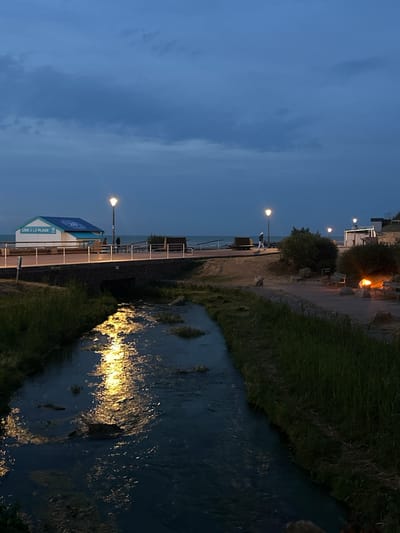 At dusk in Veules-les-Roses, France, a small river flows in the foreground, reflecting the lights of lampposts and a distant fire. The river is bordered by tall grass on the right and a stone embankment on the left, which supports a pedestrian walkway. On the walkway, a small, white building with a blue roof, labeled "LIRE À LA PLAGE," stands illuminated by a streetlamp. Further along the walkway, two figures are visible, and beyond them, more lampposts, benches, and dense shrubbery. The sky above is a deep blue, with wispy clouds, indicating the transition from day to night. The overall scene depicts a tranquil evening by the coast.