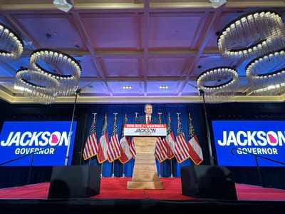 The image shows a political event likely in Atlanta, United States. An older man in a suit and red tie stands at a wooden podium on a red carpeted stage. Behind him are Georgia and American flags. A blue backdrop has “JACKSON GOVERNOR” in white letters and a pink outline of the state of Georgia. The podium also features the text “WIN BIG for GEORGIA JACKSON GOVERNOR”. Four large chandeliers are hanging from the ceiling.