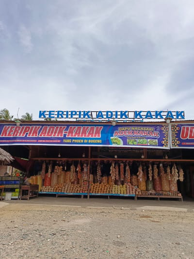 This media file captures a roadside stall in Sawang, Indonesia, selling various types of keripik (chips). The stall is adorned with banners and signs advertising "KERIPIK ADIK KAKAK" and "DAPU NAGASARI ADEK KAKAK". The signage indicates that the keripik are "Yang Phoen di Bugeng" and provides a phone number and P-IRT registration number.
The stall itself displays a wide assortment of packaged chips hanging from the ceiling and stacked on shelves and tables. The chips appear to be made from various ingredients, judging by their colors and textures, ranging from light yellow to deep orange and brown. There are also some smaller jars and containers visible, suggesting other products might be sold.
In the foreground, a gravelly, unpaved ground suggests a rural or semi-rural setting. The sky above is partly cloudy, indicating fair weather. The overall scene depicts a typical Indonesian roadside market or store, likely catering to travelers and locals looking for snacks. No people are visible in the frame. The time of day appears to be daytime, given the lighting conditions. The location is further confirmed by the text "JL. BANDA ACE", a street in Banda Aceh, Indonesia.