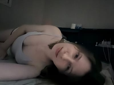 The image shows a person lying on a bed, facing the camera. The person is wearing a light-colored tank top and underwear. Their body is positioned on its side, with their head turned towards the viewer. The background appears to be a dimly lit room, possibly a bedroom, with a dark surface or furniture visible behind the person. There are some objects on this surface, including what appears to be a roll of toilet paper and some packaging. The overall lighting is low, creating shadows and a somewhat grainy texture to the image. The person's facial features are visible, and they are looking directly at the camera with a neutral expression. The location is not identifiable from the visual information in the image.
