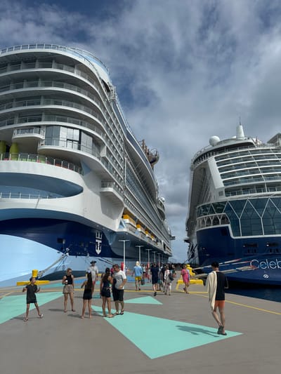 The image depicts two large cruise ships docked parallel to each other at a paved pier in the Berry Islands, Bahamas, under a partly cloudy sky.

On the left, a Royal Caribbean Group vessel, identified by its anchor logo on the light blue and white hull, features multiple decks with extensive balconies. On the right, a Celebrity Cruises ship, identified by "Celebrity" on its dark blue and white hull, also presents numerous balconies and glass superstructures. Both ships are secured to the pier with thick mooring lines.

Approximately 20-30 individuals are observed on the pier between the ships. They are dressed in casual summer attire, including shorts, t-shirts, and dresses. Actions include walking, standing in small groups, and observing the surroundings. A young male in a dark shirt and shorts stands in the lower-left foreground with arms slightly raised. The pier surface is concrete, marked with light blue geometric patterns, and features yellow bollards along the edges. Dark blue water is visible adjacent to both ships.