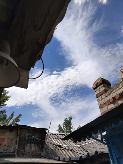 The image shows a view looking up at a cloudy sky, framed by rooftops and architectural elements. In the foreground on the left, a large, dark, overhanging structure, possibly a roof or overhang, partially obscures the view. Below it, a portion of a light fixture is visible. To the right, a chimney constructed of red and white bricks rises against the blue sky, with wispy white clouds streaking across.

In the middle ground, several corrugated metal roofs are visible, some appearing worn and aged. On the left side of the middle ground, a weathered wooden door or shutter is set into a wall. Several wires and cables run across the scene, attached to various structures. Trees with green foliage are visible peeking over the rooftops on the left and right sides. The overall impression is of an urban or semi-urban environment, with a focus on the sky and the man-made structures below. The scene is consistent with a typical residential or utilitarian area in Luhansk, Ukraine, characterized by a mix of older construction and visible infrastructure.