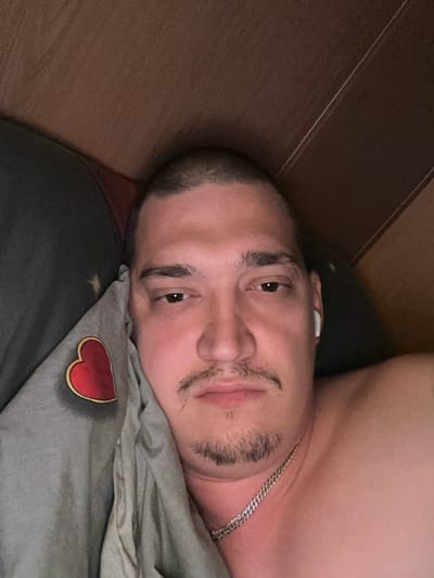 A man with short hair and a goatee is lying in bed. He is wearing a silver chain around his neck and has a white wireless earbud in his right ear. A gray pillow with a red heart design is visible on the left side of the image. The background appears to be a wooden headboard or wall. The overall setting suggests an indoor bedroom environment. The provided information does not contain details about the city of Unknown, Colombia.