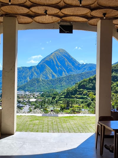 The image shows a view framed by two pillars, likely from a building, overlooking Sembalun Bumbung, Indonesia. The landscape includes a prominent mountain, lush green hills with terraced agriculture, and a town with houses and roads. A grassy area with patterned paving is visible in the foreground. Above, the ceiling has a decorative woven pattern with interspersed lights. A black electronic device is mounted on the ceiling between the pillars. A table and chairs are partially visible on the right side of the frame. The sky is blue with some clouds. The scene appears to be taken during daylight hours.