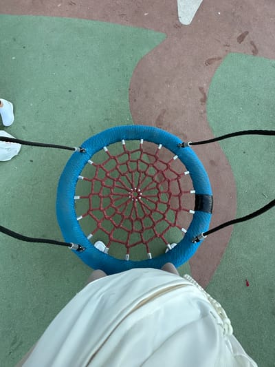 An overhead shot shows a person's legs and torso, clad in white, standing on a colorful playground surface. The person is wearing white sneakers. In front of the person is a large, circular blue swing with a red webbed net in the center. Black ropes suspend the swing. The playground surface is a mosaic of green and brown sections, with some faint footprints visible on the brown area. The scene appears to be in a public park or playground area in Sant Feliu de Guíxols, Spain.