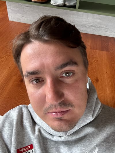 A man lies on his back with his head angled upward. He has short, dark hair, fair skin, and light brown eyes. He wears a gray hooded sweatshirt with a red name tag reading "HELLO MY NAME IS." The background consists of a wooden floor and a green cabinet with white and brown items inside.