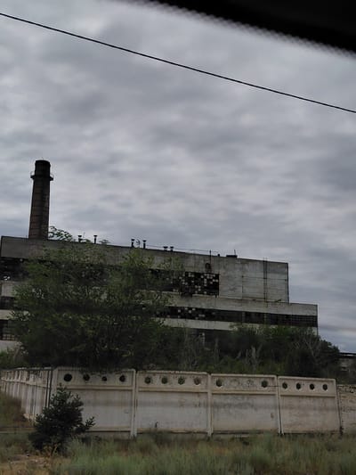 The image captures an abandoned industrial building, likely a factory or power plant, under an overcast sky. A tall, brick chimney, bearing the marking "0 19", stands prominently on the left side of the structure. The building itself is long and multi-storied, constructed from concrete and featuring numerous dark, rectangular windows, many of which appear broken or boarded up. Vegetation, including trees and bushes, partially obscures the lower levels of the building. In the foreground, a low, white concrete fence with circular openings runs across the scene, separating the industrial site from a grassy, overgrown area. The overall impression is one of disuse and decay, suggesting the industrial facility has been inactive for a considerable time. The scene is devoid of human presence or activity.