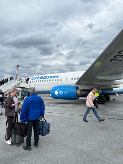 The image captures a scene on an airport tarmac under an overcast sky, likely at Bolshoye Savino, Russia. The central subject is a large passenger airplane, predominantly white with blue accents and the word "победа" (Pobeda, a Russian airline) written in blue Cyrillic letters along its fuselage. A large blue engine is visible beneath one of its wings. Part of the aircraft's registration, "73L", is visible on the underside of the wing.
Several people are present on the tarmac. In the foreground, two individuals with luggage are preparing to board or have just disembarked. One person, wearing a blue jacket, stands with a blue duffel bag on their shoulder and a grey rolling suitcase beside them. Another person, wearing a brown top and black pants with white stripes (partially visible "adidas" logo on their shoe), also has a rolling suitcase and a smaller bag. Further down, a line of passengers is visible on a mobile boarding staircase leading into the aircraft.
Near the airplane's engine, a person in a light pink jacket, jeans, and a yellow safety vest stands on the tarmac, possibly ground crew or another passenger waiting. The ground is asphalt, and various pieces of airport ground equipment, such as a small vehicle near the boarding stairs and possibly another truck further back, are present. The sky is filled with grey, heavy clouds, indicating a cloudy or overcast day. The overall activity suggests passengers are in the process of boarding or disembarking the Pobeda airline flight.