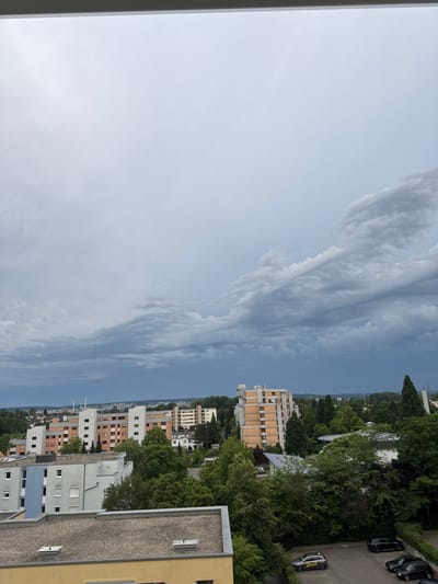 The image shows an overcast sky with dramatic cloud formations over a residential area in Sindelfingen, Germany. In the foreground, the rooftops of several apartment buildings are visible, constructed with concrete and gravel surfaces. One building in the lower left corner has a light blue facade, while another adjacent building is gray. Further into the scene, several taller apartment buildings with white and orange exteriors stand amidst lush green trees. A few cars are parked on a paved area in the lower right of the image. The background shows a cityscape with more buildings and industrial structures visible under the heavy clouds.