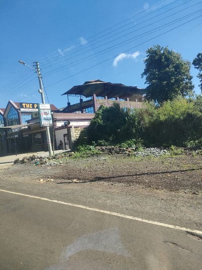 The image shows a view of a roadside scene in a municipality ward in Kenya. The sky is clear blue with a few wispy clouds. In the foreground, there is a paved road with a white line marking. To the left of the road, there is a gravelly verge. Beyond the verge, there is a building identified by a sign as "THE P". A lamppost stands near the building. Adjacent to "THE P", there is another building with a covered rooftop area. The buildings sit slightly uphill from the road, with vegetation and rocks covering the slope. A person is seated near the entrance of the second building. Power lines run across the upper portion of the image.