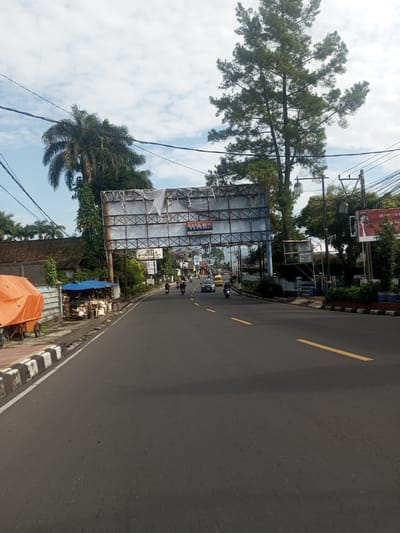 The image shows a street in a tropical location. A large, aged billboard spans the road. The road itself is asphalt with yellow lane markings. Cars and motorbikes are visible in the distance. Trees line the street, including palms and a tall conifer. There's a small vendor stall to the left with blue tarpaulin. Electrical wires cross overhead. The sky is partly cloudy. The scene does not obviously align with Zeyathiri District, Myanmar based on architectural style, common vegetation, and general visual characteristics. The information about location may be inaccurate.