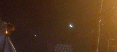 The image shows a dark night scene in what is possibly Ndenderu ward, Kenya. A crescent moon is visible in the upper right quadrant. A lit street lamp appears on the left side. The blurred text "ROYAL PRAIRIE SCHOOL" is visible, possibly a sign for a nearby institution. A tall, thin, partially lit vertical structure on the right-hand side may be a utility pole or similar infrastructure. A bright spot is visible under the moon, possibly a star or another light source.