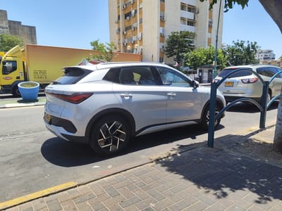 In Kiryat Yam, Israel, on a sunny day, a white BYD Atto 3 electric vehicle is parked on the right side of a two-lane asphalt road. The car's rear is facing the viewer, and its distinctive geometric wheel design is visible. To the left of the BYD, a beige SUV is also parked. Further to the left, a bright yellow truck with a large cargo box is stopped, with a blue planter containing green vegetation situated in front of it. The truck has Hebrew text on its side, indicating it belongs to "DON" and includes a phone number. In the background, a tall, multi-story apartment building with numerous air conditioning units and satellite dishes is prominent. Trees and a clear blue sky complete the urban street scene.