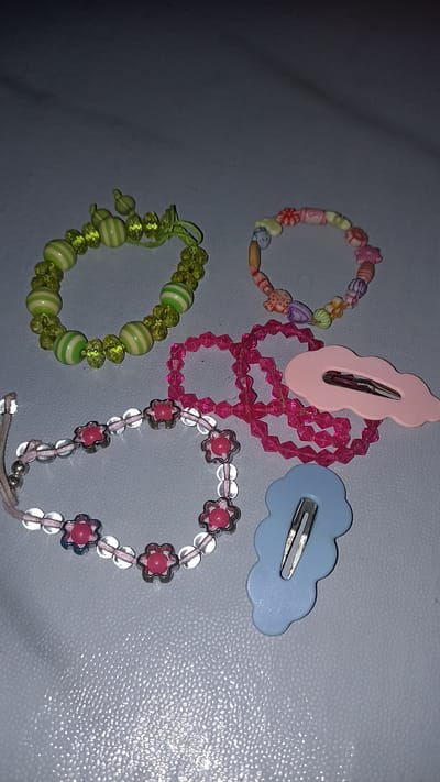 This is a close-up, eye-level shot of several colorful beaded bracelets and two hair clips arranged on a textured, light-colored surface, possibly a table or countertop. The lighting is dim, creating shadows and highlighting the glossy surfaces of the beads and clips.
In the upper left is a bracelet made of alternating light green and white striped beads, interspersed with faceted clear green beads. Next to it, to the right, is another bracelet with small, colorful beads resembling candies and flowers in shades of pink, orange, yellow, and blue.
Below these, a bright pink string of faceted beads forms a square shape, with a few loose beads scattered around it. To the right of the pink beads is a light pink, cloud-shaped hair clip with a silver metal clasp.
In the lower left, a bracelet features clear beads and small, pink, flower-shaped charms with silver metal accents. Adjacent to it, to the lower right, is a light blue, cloud-shaped hair clip with a silver metal clasp.
The scene is entirely indoors and does not provide any specific geographical context for the city of Sedayu, Indonesia. There are no people, actions, or interactions depicted in the image.