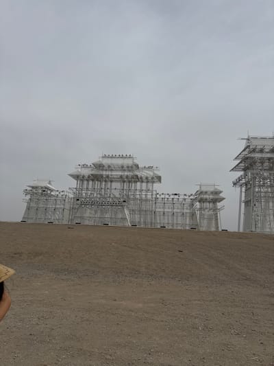 The image shows a large-scale architectural art installation on display outdoors in Guazhou County, China. The installation consists of multiple pagoda-like structures constructed from metal scaffolding, creating a complex, linear cityscape silhouette. The sky is overcast and gray, suggesting a muted, diffused light. The ground is dirt or gravel, with sparse vegetation. The installation appears to stretch away into the distance along the ground, following the slight decline. A blurred partial view of a person is in the bottom right corner. The architecture appears light and airy, contrasting with the stark background of sky and ground.
