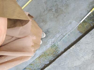 A person wearing a light brown hijab and light-colored sneakers is standing next to a shallow, narrow concrete trench filled with flowing water. A yellow and white hose is visible at the upper right of the image, with water being sprayed from it into the trench. The ground around the trench is concrete and appears wet in places, with some dirt and debris. The image is taken from a high angle, looking down at the scene. The location is identified as Suzak, Kyrgyzstan.