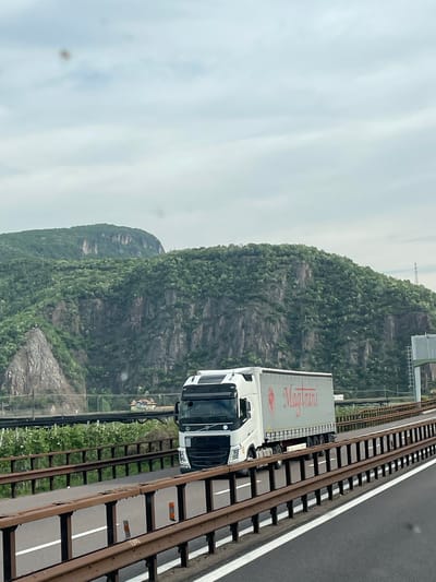 A slightly tilted outdoor shot depicts a white semi-trailer truck, with the name "Mag Trans" visible on the trailer, moving along a highway beside a railway track. The truck is positioned to the right of the frame. To the left, a mountainous landscape rises, showing a rocky terrain covered in greenery. The sky is overcast, creating a diffused lighting effect. The highway and railway track are both parallel and run along the bottom of the frame, with the railway track closer to the camera. The location is possibly in or near Auer - Ora, Italy.