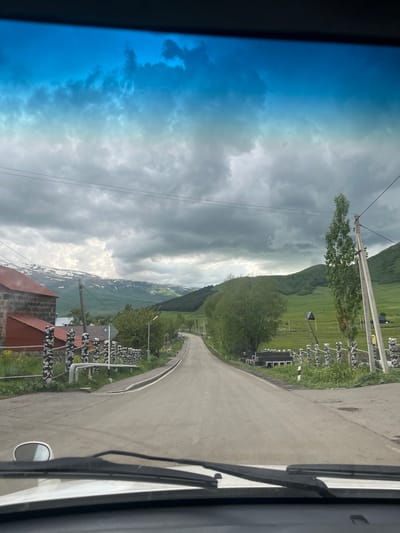 The image captures a view from inside a vehicle, looking out through the windshield. The scene depicts a rural road in Lusagyugh, Armenia. The road, paved with asphalt, curves gently into the distance, flanked by green rolling hills and mountains in the background. The mountains are capped with patches of snow, indicating a high altitude or a recent snowfall. The sky is partly cloudy, with dramatic grey clouds dominating the upper portion of the frame, interspersed with brighter patches suggesting sunlight breaking through.
On the left side of the road, there is a stone building with a red roof, characteristic of rural Armenian architecture. Several fences, constructed from stacked stones and topped with what appear to be rough-hewn logs or branches, line the roadside. These fences create a rustic boundary. A tall, slender tree stands prominently on the right side of the road, near a concrete utility pole with overhead wires. Further down the road, more houses and rural structures can be seen nestled amidst the greenery. The overall impression is one of a serene, mountainous countryside, likely experienced during a drive through the village.