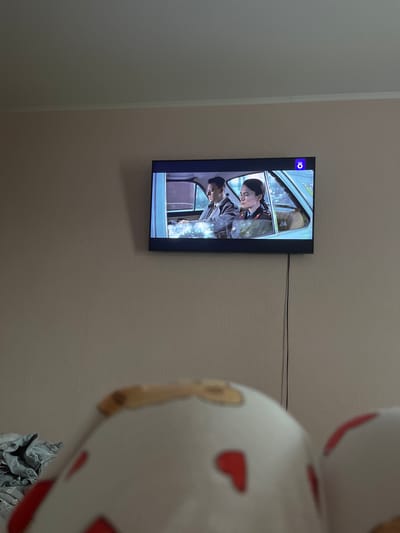 A television screen mounted on a wall displays a scene from a film or show. Two individuals are visible inside a light blue car. The person in the driver's seat, a man wearing a suit, is looking ahead. The passenger, a woman in a military-style uniform, is also looking forward. A blue logo with white circles and a white 'O' is present in the upper right corner of the screen. A black cable hangs down from the television to the wall. The wall appears to be painted a light tan color, and a white crown molding is visible near the ceiling. In the foreground, out of focus, are two large, white objects with red heart-shaped markings, suggesting they might be decorative pillows or toys. The overall environment appears to be an indoor living space. The image was taken in Sof'ino, Russia.