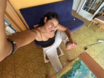 An indoor wide-angle selfie depicts an adult female seated in a residential room located in Vilanova I La Geltrú, Spain. The precise address or specific area within the city is not discernible from the image.
The subject is an adult female with medium-toned skin and dark, wavy hair pulled back, visible on the left side of the frame. She is wearing a black spaghetti-strap crop top and white wide-leg trousers. Her left arm is extended towards the camera, holding the device used to capture the image; multiple dark-colored bracelets are visible on her left wrist. She is smiling with squinted eyes and a slightly open mouth, looking directly at the camera. She is seated on a chair with a dark blue cushion, part of which is visible behind her. Her left foot is resting atop her right thigh, which is extended forward, with her right foot resting on a small, light brown stool.
The floor is covered with yellowish-brown ceramic tiles featuring a repeating geometric pattern of squares and diamonds. In the background, a large dark blue folded futon or sofa is positioned against a solid yellow wall. To the immediate left, a tall, light brown wooden bookshelf packed with books and binders is visible. To the right, a smaller light brown wooden bookshelf is partially visible, also containing books, with a light-colored cushioned chair or stool adjacent to it. A brown wooden desk occupies the lower right foreground, topped with a mousepad displaying an impressionistic landscape painting, reminiscent of Van Gogh's style. A white power strip with black electrical cords emanating from it lies on the tiled floor near the desk. A white cable is also visible resting on the dark blue futon.