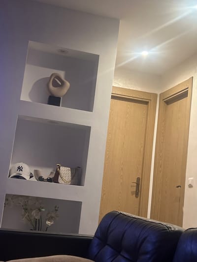 The image depicts a low view of an interior space featuring a built-in shelving unit with three square compartments. The left compartment holds a light brown sculptural object beside a black rectangular object. The middle compartment displays a handbag, sunglasses, and a baseball cap. The right compartment contains a floral arrangement with white flowers. Above the shelving unit, two wooden doors are visible, with the upper door appearing to be an access panel. Part of a dark blue leather sofa is visible on the right edge of the image.