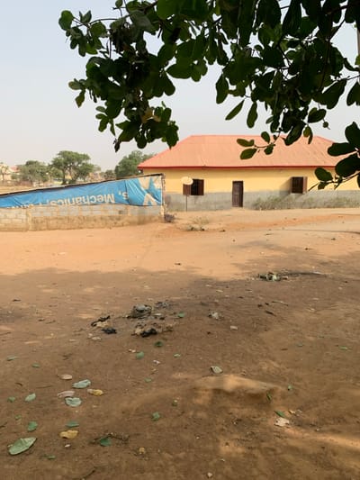 The scene is an outdoor, daytime view in Bwari, Nigeria, dominated by a dirt ground and architectural structures.
In the foreground, large, dark green leaves from an overhead tree partially obscure the upper portion of the frame. The immediate ground consists of reddish-brown dirt, uneven and sparsely covered with scattered dried leaves and small pieces of litter, including dark plastic and unidentified debris. A small, dark, irregular area of disturbed earth or burnt material is visible centrally on the ground.
To the right, a single-story building with light yellow walls and a corrugated orange-red metal roof stands. It features three rectangular windows with dark frames and security bars. A single dark brown wooden door is centered on its facade. A small, circular, dark-colored satellite dish is mounted on a pole to the left of the rightmost window. The lower section of the building's walls is a darker grey.
To the left of the building, a low wall constructed of grey concrete blocks is present. A large, faded blue tarp, bearing partially inverted white text that reads "S. Mechanics," covers the upper section of this wall, extending upwards.
The background shows a pale, hazy blue sky. Distant green trees and faint outlines of other light-colored buildings are visible on the horizon under the hazy sky. No individuals are present in the image, and no active events are depicted.