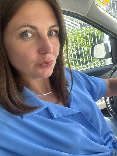 A woman with brown hair and blue eyes is in the driver's seat of a car, looking at the camera. She is wearing a blue collared shirt and a silver necklace. Her lips are pursed. Outside the car window, a metal fence and green foliage are visible. There is a sticker on the inside of the car's windshield. The image is taken from a medium close-up perspective, focusing on the woman's face and upper body. The scene is outdoors during the day, with sunlight illuminating the environment. The car appears to be parked or stopped, given the static nature of the composition.