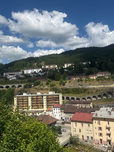 The image depicts a view of the city of Hauts de Bienne, France, with a focus on its urban landscape nestled against a forested hillside. Several multi-story residential buildings are visible, constructed in a mix of architectural styles and colors, ranging from pale yellow and white to blue and brown. Some buildings feature balconies and numerous windows, suggesting apartments or condominiums.
The city appears to be built on a series of terraces, with roads and pathways winding through the slopes. A prominent arched viaduct, likely a railway bridge or road structure, spans across the lower part of the scene, with cars visible on the road beneath it. Further up the hillside, more buildings are scattered amongst the trees, and evidence of terraced landscaping can be seen.
The surrounding environment is dominated by a dense, dark green forest covering the steep hillside that rises behind the city. The sky above is a bright blue, dotted with fluffy white cumulus clouds, indicating fair weather. The foreground features lush green foliage, suggesting a verdant natural setting around the urban area. The overall impression is of a town integrated with its natural, hilly environment under a sunny sky.