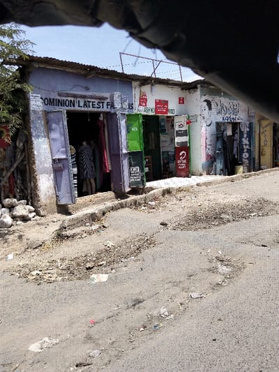 This image displays a street scene in Mkomani ward, Kenya, featuring a row of single-story commercial buildings under clear daylight. The perspective is slightly elevated, with a dark, irregular object partially obscuring the top of the frame.
From left to right, the observed structures include:
1. A light purple-grey building with a corrugated metal roof. Its entrance, comprising two open metal doors, reveals a dark interior with clothing items and a mannequin wearing a striped dress. A sign above the entrance reads "DOMINION LATEST FASHIONS." To the left, a small tree or bush and a pile of rocks are visible.
2. An adjacent white-painted building, also with a corrugated metal roof. Its facade is covered with various red and white posters and signs, notably indicating "M-PESA" services and "ASH NO RAISES." A green KCB-branded ATM-like machine and a red-and-white 'Airtel Money Agent' stand are positioned at its entrance.
3. A contiguous white-painted building with graffiti on its exterior, including a stylized facial depiction. Its doorway appears dark.
4. A partial section of a yellow-painted building is visible on the far right.
The asphalt street in the foreground shows significant deterioration, characterized by multiple cracks, potholes, and areas of loose dirt and gravel. A concrete curb separates the roadway from the building fronts. Small pieces of debris and litter are scattered across the street surface. No individuals are visibly present, with the exception of mannequins inside the "DOMINION LATEST FASHIONS" store.