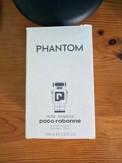 A white rectangular box, likely packaging for a fragrance, is the central subject, positioned on a light brown wooden surface. The box is oriented vertically within the image frame. On the left side of the box, the word "PHANTOM" is printed vertically in large, dark gray capital letters. On the right side, a stylized robot graphic featuring a 'P' within its design is visible. Below this graphic, the text "TESTER - PROBADOR" is printed, followed by "paco rabanne". Further details include "EAU DE TOILETTE", "NATURAL SPRAY", and "100 ml e 3.4 FL OZ". The wooden background displays prominent grain patterns and several dark knots. A partial view of a dark, curved object, possibly a cushion or part of a chair, is visible on the far left. The image presents a static product shot of the fragrance box. The item is located in Saedinenie, Bulgaria.
