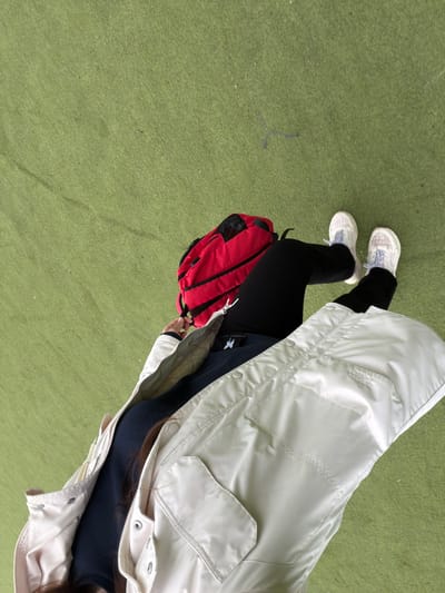 The image shows an overhead view of a person standing on a green surface. The person is wearing black pants and white sneakers, and a long, light-colored coat. A red backpack is slung over their shoulder. The green surface appears to be artificial turf or a similar material. The location is unspecified, therefore, the reference to Tokkari-Lend, Russia, is not relevant.