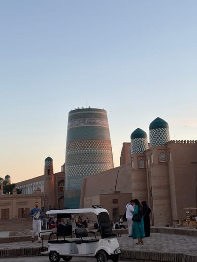 In the historic city of Khiva, Uzbekistan, a wide-angle, eye-level outdoor shot captures a scene in the Itchan Kala, the walled inner city. The sky is a clear, light blue, with a hint of sunset warmth on the horizon. Prominently featured is the Kalta Minor minaret, a distinctive teal and patterned cylindrical tower, to the left of the frame. To its right, a section of the ancient city walls with crenellations and two teal-domed structures, possibly minarets or towers, rise against the sky. The walls are constructed of weathered brick, typical of Khiva's architecture.

In the foreground, a white electric cart, commonly used for tourism within the Itchan Kala, is parked. A man wearing a blue shirt and white pants walks past the cart, holding a bag. To the right of the cart, three individuals are gathered, engaged in conversation: a man in a white shirt, a woman in a teal dress, and another woman in dark attire. Behind them, other tourists are visible in the distance, as well as more traditional brick buildings with arched windows. The ground surface is paved with cobblestones. The overall impression is one of a serene, late afternoon in a culturally rich historical site.