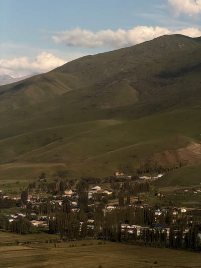 The image depicts a serene mountain landscape with a small village nestled in the valley. The foreground is dominated by a steep, grassy mountain slope that stretches diagonally across the frame. The texture of the grass varies, with some areas appearing lush and green, while others are drier and more brown. Several distinct ridges and curves are visible on the mountainside, creating a sense of depth and scale.
In the background, a cluster of houses forming a village is scattered among a dense line of tall, slender trees. The houses are of various colors and architectural styles, with some featuring white walls and contrasting roofs. The trees are densely packed, creating a natural screen that partially obscures the village. The overall impression is one of a rural, perhaps isolated, settlement.
To the left of the mountain, a patch of blue sky is visible, dotted with soft, white clouds. The lighting suggests it is daytime, likely late afternoon or early evening, given the soft shadows and warm tones in the grass and village. The weather appears clear with some scattered clouds. The location cues, such as the mountainous terrain, the style of the village, and the vegetation, are consistent with a rural setting in a mountainous region like Karagai Bulak, Kyrgyzstan. No people are explicitly visible, and the scene conveys a sense of tranquility and natural beauty. No text is visible in the image.