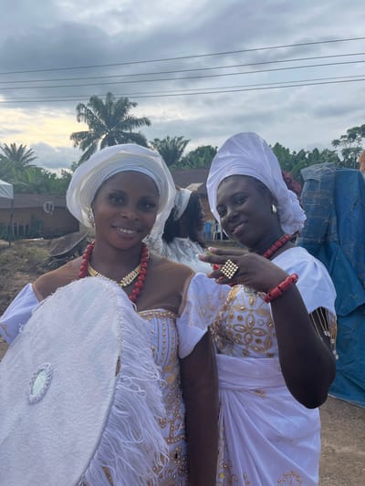This image captures two women in traditional attire, smiling and posing for a photograph outdoors in Aghalokpe, Nigeria. Both subjects are dressed in elegant white garments, featuring intricately embroidered gold patterns on their dresses and matching white headwraps (gele).

The woman in the foreground wears a multi-strand red coral bead necklace with gold details, a red coral bead bracelet, and a prominent square-patterned ring on her right hand. She holds a large, round white accessory, possibly a feathered fan or ceremonial object, which has white fringes around its edge and a small mirror embedded in its center. The woman positioned slightly behind her is similarly adorned with a red coral bead necklace and bracelet, and a large ring. Both women are smiling broadly, conveying a sense of happiness and warmth.

The setting appears to be an outdoor gathering or celebration, possibly under an overcast sky, as indicated by the cloudy background. Tall palm trees are visible in the distance, characteristic of the tropical landscape in Nigeria. Power lines traverse the upper portion of the sky, suggesting an area with some infrastructure. The lighting suggests it is daytime, possibly late afternoon given the soft quality of light. No discernible text is visible.