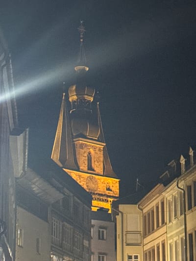 This is a nighttime, low-light, medium-shot photograph of the Wendelinusbasilika (St. Wendelin's Basilica) in Sankt Wendel, Germany. The tower of the basilica, with its distinctive stepped roof and spire, is illuminated by artificial lights, casting a warm yellow glow. The tower features a clock face visible on its side. In the foreground and to the sides of the frame, buildings with darkened facades and visible windows are present, suggesting a narrow street or plaza. A bright beam of light cuts diagonally across the sky from the left side of the image, likely from a spotlight or other external light source. The sky is dark, indicating it is nighttime. There are no people visible in the image. The overall scene depicts an architectural landmark at night, surrounded by urban buildings.