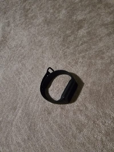 The image displays a single black wrist-worn device, likely a fitness tracker or smartwatch, resting on a light grey, textured surface consistent with a carpet or rug. The device is oriented diagonally from the top-left to the bottom-right of the frame, with its rectangular display unit visible. A shadow is cast from the device towards the bottom-right, indicating a light source from the upper-left. No individuals, other objects, or discernible actions are present in the scene, which is located in the city of Cholakova, Bulgaria.