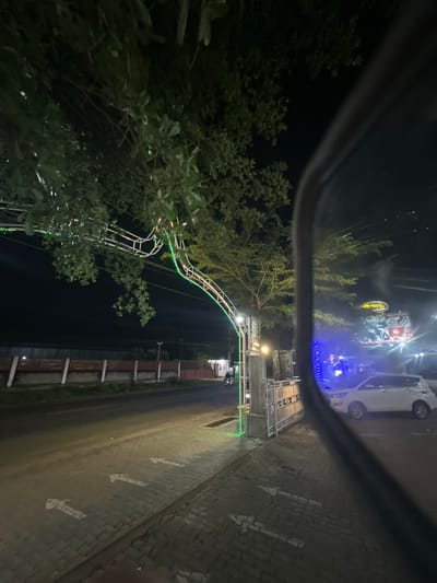The image depicts a nighttime street scene in Keudeteupinpunti, Indonesia, as viewed through a car's side mirror. The foreground is dominated by the dark rim of the mirror, obscuring part of the view. Reflected in the mirror is a white vehicle parked on the right side of the frame.
Beyond the mirror's reflection, the scene shows a paved road with white directional arrows painted on it. To the left of the road, a pedestrian walkway is visible, made of interlocking bricks. A decorative archway with green lights spans across the road, illuminated by a warm-toned streetlamp at its base. Trees with dense foliage hang over the archway.
Further down the street, a low fence with horizontal planks and vertical white posts runs along the side. In the background, beyond the fence, are buildings and the silhouette of trees against the night sky. A large, illuminated billboard is visible to the right, with text and images that are difficult to discern clearly due to the distance and lighting. The overall atmosphere is that of a commercial or residential area at night, with festive lighting on the archway suggesting it might be a business entrance or a decorative street feature.