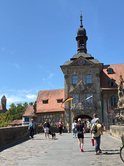 The image depicts a sunny day scene on a cobblestone bridge in Bamberg, Germany, likely the Obere Brücke. In the foreground, several people are walking on the bridge. An older man in a blue vest and dark pants walks towards the viewer on the left. To his right, a woman with a white skirt and black top walks with a small brown dog. Behind them, other pedestrians are visible, including a group of people walking away from the camera.
Dominating the background is a distinctive building, the Alte Rathaus (Old Town Hall), characterized by its ornate facade, a clock face with Roman numerals, and a tower with a bell. Two flags, the German flag and another blue and white flag, fly from a balcony. To the left of the town hall, a distant church tower is visible. The bridge itself is lined with a stone wall, and a fence separates the bridge from an ornate statue on the right. Lush green trees are visible behind the buildings on the left. The sky is a clear, bright blue with a few wisps of clouds.