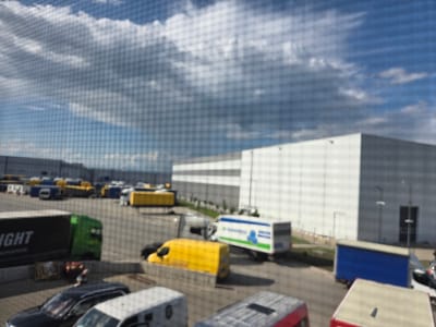 The image, captured through a fine mesh screen, shows a busy industrial or logistics park in Bozhurishte, Bulgaria, under a partly cloudy sky. Large, modern warehouse buildings with white and grey facades form the backdrop. The extensive paved area in the foreground and midground is filled with numerous commercial vehicles.
These vehicles include several delivery trucks and vans: a green-cab semi-truck with a black trailer displaying partial text "IGHT NOVATION" (likely part of "BRIGHT INNOVATION"), various yellow-bodied vans, and a white box truck with the branding "TRANSPRESS" visible in blue and green. A blue box truck and several smaller cars and white vans are also present. One person is vaguely visible standing near a dark car in the lower-left section of the image. The time of day is daytime, with bright natural light creating visible shadows. The weather is partly cloudy, featuring white and grey clouds against a clear blue sky. The scene depicts routine operations at a commercial distribution or shipping hub.