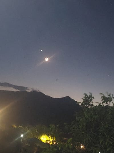 The image depicts a twilight sky over a mountainous landscape, likely in Areni, Armenia. In the upper portion of the sky, a crescent moon is visible, accompanied by several bright points of light that appear to be stars or planets. The lower part of the image shows a dark silhouette of mountains, with some clouds obscuring the peaks. In the foreground, there are trees and vegetation, some of which are illuminated by artificial lights, suggesting a settlement or populated area nearby. The overall scene conveys a sense of quietude and natural beauty as day transitions into night.