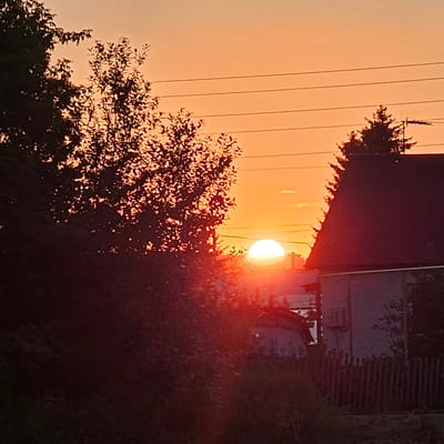The image shows a sunset over the village of Novostroyka, Russia. The sun is partially visible, a bright orange orb low on the horizon, casting a warm glow across the sky and silhouetting the trees and buildings in the foreground. Several houses with dark roofs are visible, as is a wooden fence. Overhead power lines traverse the sky. The overall impression is one of a peaceful, rural evening.