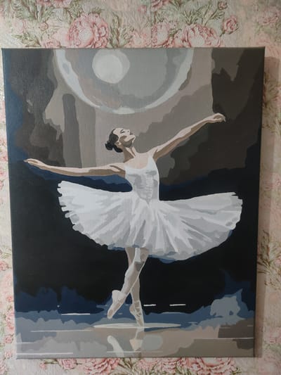 A painting depicts a ballet dancer in a white tutu and bodice, poised en pointe with arms extended. The dancer's head is tilted back, and her eyes are closed. The background is abstract, with shades of gray, blue, and brown, suggesting a stage or a moonlit sky. The painting is displayed against a floral-patterned wallpaper. The location is identified as Ternovka, Moldova.