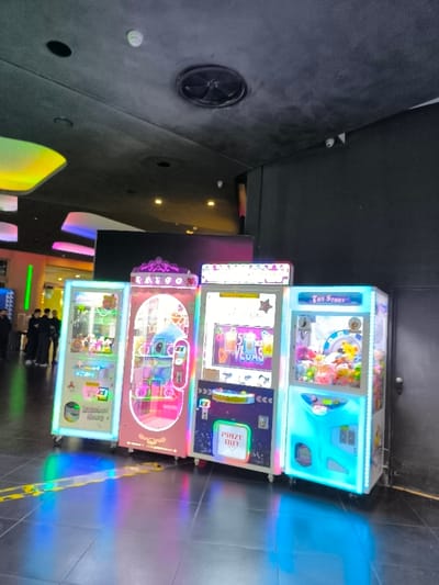 The image displays an indoor scene in Agios Dimitrios, Greece, featuring a row of four brightly illuminated arcade prize machines. These machines, likely claw cranes, emit vibrant LED lights that cast colorful reflections onto the dark, polished floor.

From left to right, the machines exhibit distinct designs: the leftmost is predominantly light blue with the text "Rainbow Story"; next is a pink machine labeled "KAIGO"; the third machine is dark blue/purple with "5 VEGAS" and "PRIZE OUT" displayed; and the rightmost machine is light blue and branded "Toy Story." Each machine is filled with various plush toys and small prizes.

In the blurred background to the left, several indistinct human figures can be seen, indicating a public area. The ceiling is dark grey/black, featuring recessed lighting, a white smoke detector, a circular ventilation grille, and a small security camera mounted near the wall-ceiling junction on the right.

The overall setting suggests a modern entertainment center, a shopping mall, or a cinema lobby. The bright, artificial lighting creates an engaging and playful atmosphere. No specific time of day or weather conditions can be inferred from the indoor environment.