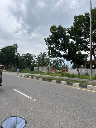 A man walks on the side of a road in Tanahterban, Indonesia. He is wearing a light blue short-sleeved shirt and light-colored shorts. The road is paved and has a white line marking the lane. The man is walking away from the viewer, towards the right side of the frame. The area is lined with trees, some of which are palm trees. In the background, there are buildings and construction equipment, including a yellow excavator. The sky is overcast with gray clouds. The image appears to have been taken from a moving vehicle, possibly a motorcycle, as a portion of a side mirror is visible in the lower left corner of the frame.