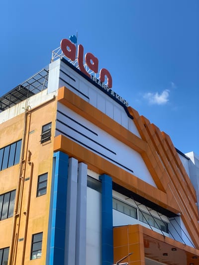 The image shows the exterior of a building in Sungai Paring, Indonesia. The building has an orange facade with blue and white accents. A sign at the top of the building reads "alan" in large red letters, with "PRIMARY & KIDS" written below in smaller letters. The sky is blue with a few scattered clouds. The building appears to be a commercial establishment, possibly a school or a children's center. The image is taken from a low angle, looking up at the building. There are no people visible in the image.