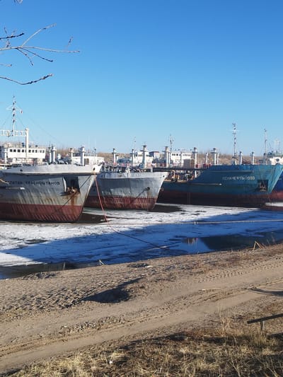 The image shows several ships docked in the city of Zhatay, Russia. The ships are partially submerged in a body of water with patches of ice. The shore is sandy with sparse vegetation. The sky is clear and blue. The ships are of varying sizes and colors, with the names "KAPITAN KRASNOSHTANOV" and "LENANEFTE-2055" visible on two of them.