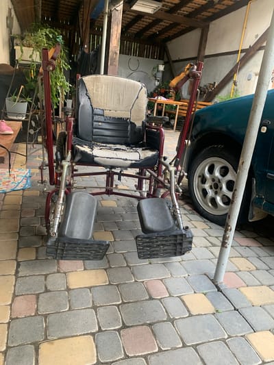 The image shows a red metal-framed wheelchair, likely a custom-built or modified one, positioned on a paver patio in an outdoor setting. The wheelchair features two padded seats, one in black and beige, and two dark grey footrests with a ribbed texture. The overall structure of the wheelchair suggests it might be designed for maneuvering over uneven terrain or perhaps for use with a vehicle.
In the background, part of a dark teal car is visible, specifically its rear wheel and a portion of the chassis, suggesting the wheelchair is parked near a car. There's also a wooden structure with beams and siding, possibly a shed or carport, providing some overhead cover. Green plants, including what appears to be a Christmas cactus, are present in pots, indicating a cultivated outdoor space. The ground is paved with interlocking concrete pavers in various shades of grey, yellow, and pink.
The scene appears to be outdoors during the daytime, with natural lighting. There is no discernible activity taking place; the wheelchair is stationary. The overall impression is one of a somewhat cluttered but functional outdoor area, possibly a backyard or a workshop space. No people are visible in the image. The time of day is difficult to determine precisely, but the lighting suggests it's not early morning or late evening. The location cues, such as the paver patio and the style of the wooden structure, are consistent with residential or rural Ukrainian settings.