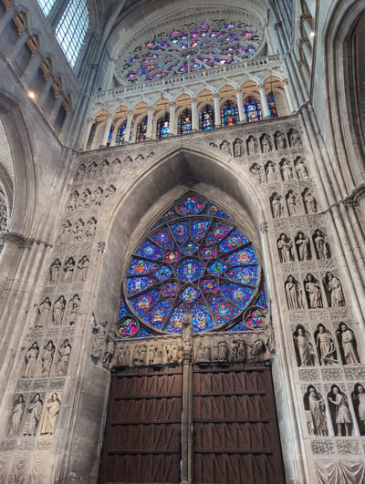 The image shows a view of a grand cathedral interior, with a large rose window dominating the center of the frame. The rose window is made of stained glass and features intricate patterns in shades of blue, red, and purple. Below the rose window are massive wooden doors, ornately carved and set within a stone archway. The walls of the cathedral are adorned with numerous stone sculptures of figures, arranged in rows within arched niches. The architecture is Gothic, characterized by pointed arches, ribbed vaults, and soaring ceilings, with sunlight filtering through the stained glass, casting colorful patterns on the stone. This is the interior of the Cathédrale Notre-Dame de Reims, located in Reims, France, not Urvillers, France.